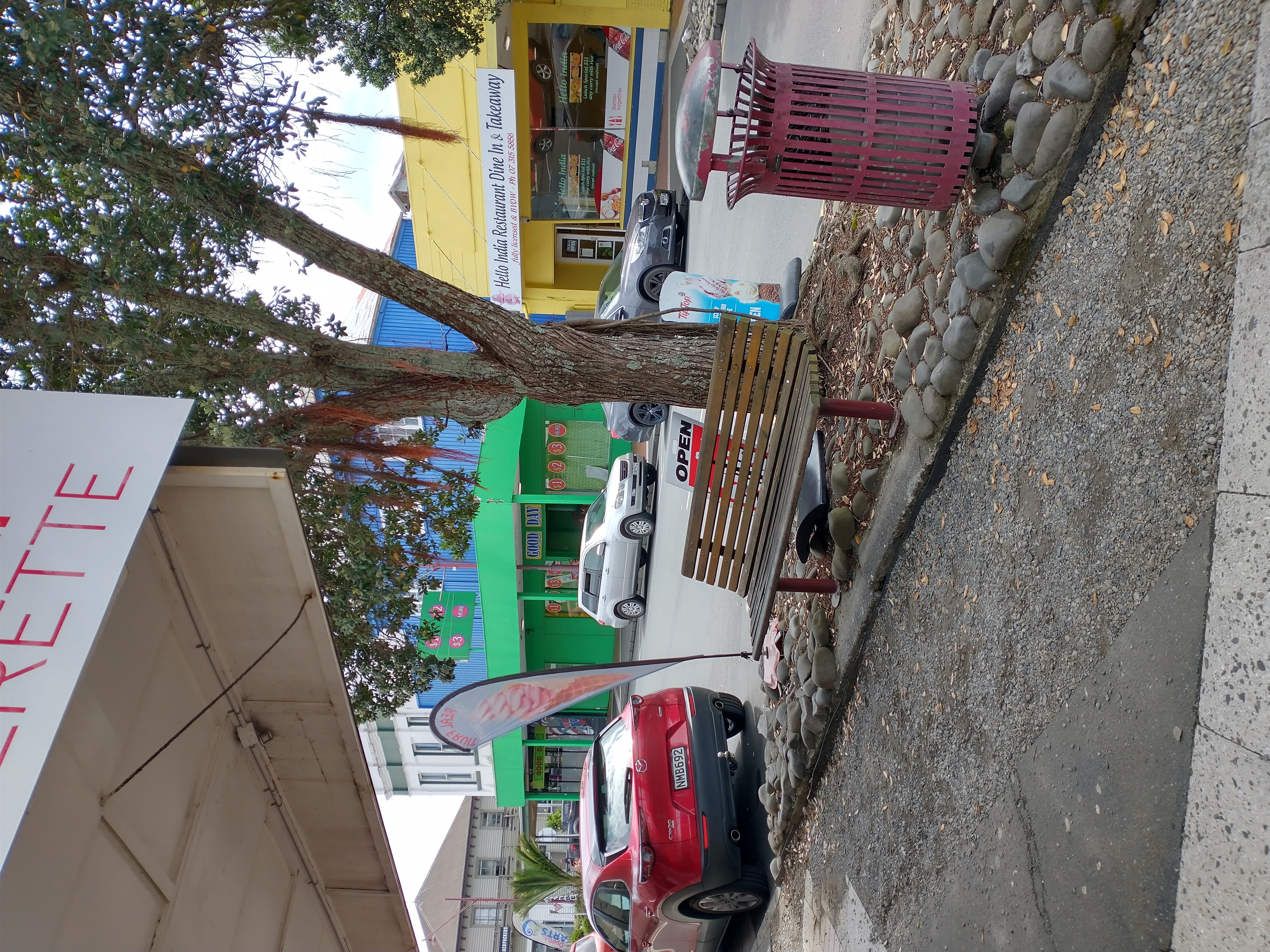 A photo of a section of footpath on Church Street Opotiki showing where work has been done. Includes cars parked next to footpath, a park bench under a tree and shops.